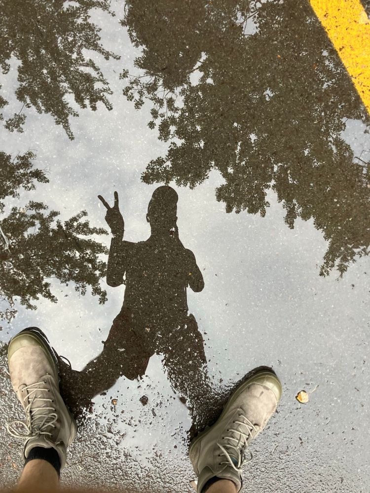 someone taking a selfie by leaning over a puddle in the street. the figure is silhouetted in the puddle of water. their boots are visible at the bottom of the frame where their feet meet the reflection's. there is a yellow stripe of street paint at the top right. there are tree branches with leaves visible in the background behind them.