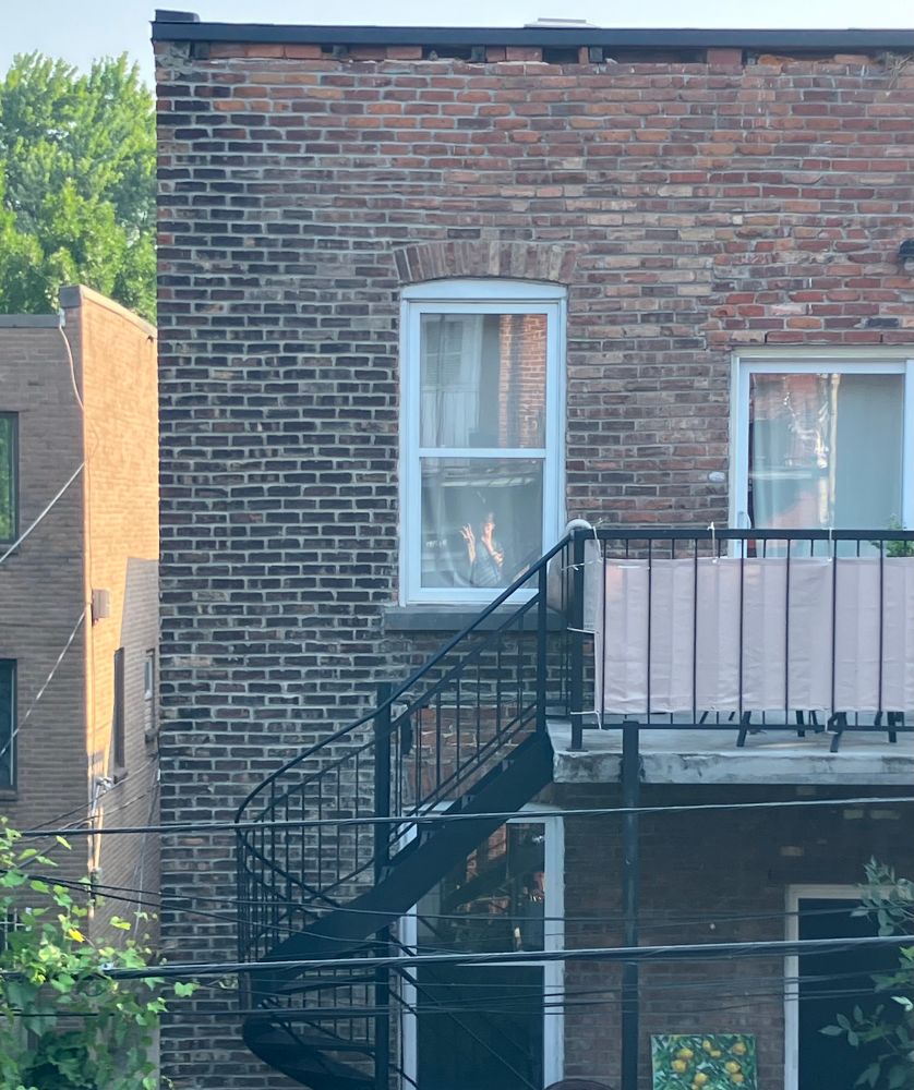 picture of the back of a three story apartment building around sunrise. there is a winding black metal staircase leading up to a balcony. the balcony has a black metal railing with pink fabric stretched between the bars. the building has a brick facade, with white-framed windows. in the center window is visible the distant reflection of the person taking the photo