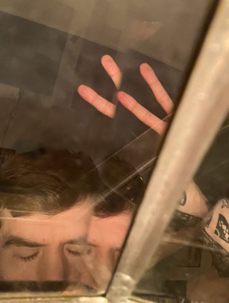 a person's face reflected through glass. there are multiple panes of glass at weird angles to each other, creating multiple reflections of the person and his hand. he's holding up a peace sign. there are black tattoos visible on his raised hand. his face is only visible from the middle of his nose up. he has brown slightly wavey short hair. his eyes are closed. a metal bar or seam between panes of glass runs from the bottom of the frame to the top right.