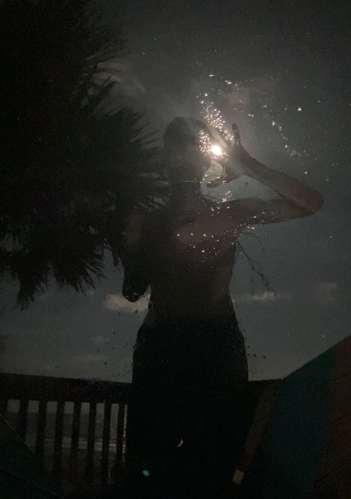 selfie of a person dimly lit, in silhouette, by a bright light from behind. they are holding up their hand to their head, with the bright light visible between their fingers. there is a palm tree visible on the left side of frame, above a low railing. there is water on whatever surface the person is taking the reflected selfie in, making little drop of illumination across its surface. there are a few clouds visible behind the dark figure