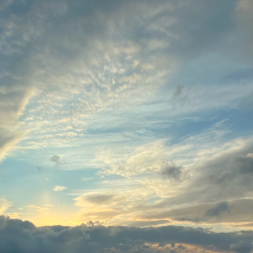 a complex arrangement of clouds over a blue sunset-lit sky. there are various sections of fluffy white clouds, other clouds that are closer and less illuminated (so they are darker), and another darker band of clouds across the bottom of the frame