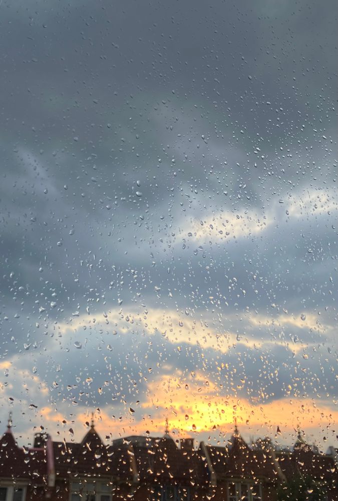 a row of roofs of houses stretches across the bottom of the frame. they are out of focus. there is a pane of glass completely covering the field of view, and on it are wet droplets. the sun is setting in a cloudy sky. the clouds grow darker and greyer towards the top of the image