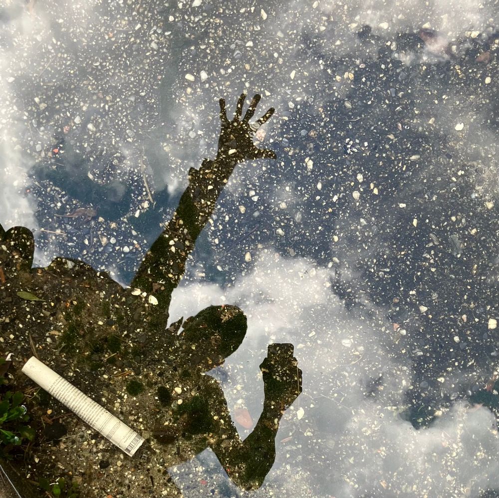 a reflection selfie. a shadowy figure is reflected in water from above. they are sticking their hand out into the air above them, with fingers splayed out. they are holding their phone out to take the selfie. behind them in the sky above there are big fluffy clouds. below the surface of the water you can see the grit of gravel. an old rolled up newspaper rests on the watery floor inside the person's silhouette
