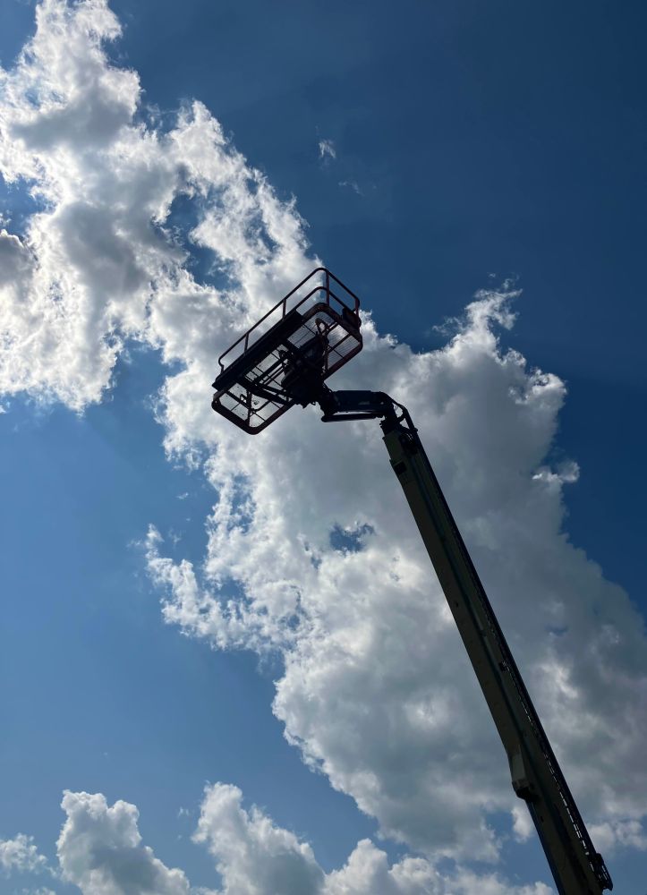 a picture of a boom lift from below. the boom begins at the bottom right of frame and stretches towards the opposite top left corner. the lift platform is roughly two thirds of the way towards that corner. behind the boom lift there is a long fluffy stretch of clouds, in a diagonal that runs parallel to the boom. the sun is shining through the clouds from behind