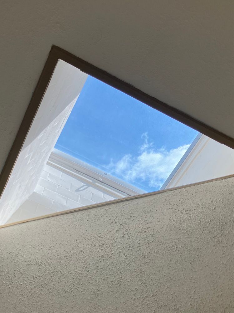 looking up through a skylight that has been cut diagonally through a wall. it creates a triangular portal with a view of the sky. the walls are white with a brown wooden trim. there is a single solitary cloud moving lazily across the frame.