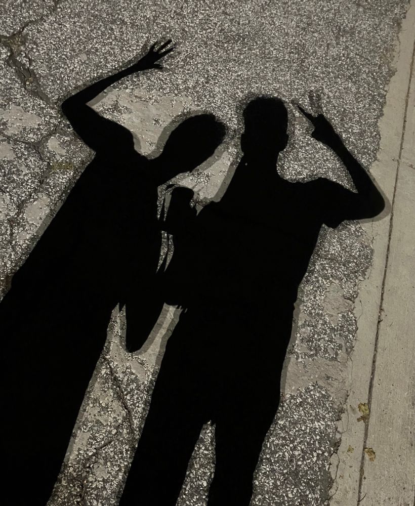 the shadow of two men against the pavement at night. the shadows are actually made from multiple overlapping light sources creating a multilayered pattern to the shadow edges. the right shadow is holding up his left hand in greeting. in his left hand he is holding a cup. the right shadow is holding up a peace sign