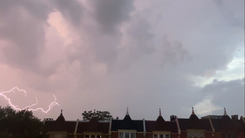 a long row of house rooftops stretches across the bottom of the photo. the rooves have weird little ornaments at the top of them. they look like little wooden hookahs while at the same time appearing completely innocuous and plain in the context. at the left side there is a treetop visible. Most of the frame is taken up by clouds. the clouds are darker towards the top left of the frame, and lighter to the bottom right, where a bit of blueish sky is visible in patches. at the left bottom of the image, above the treetop, there is a jagged white lash of lightnight across a portion of the sky