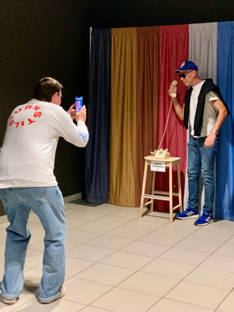 Im Hintergrund ein Vorhang mit dicken bunten Streifen. Davor posiert ein Mann mit einem Fake-Telefon. Im Vordergrund steht ein Mann in einem weißen Turnstile-T-Shirt der davon ein Foto macht 