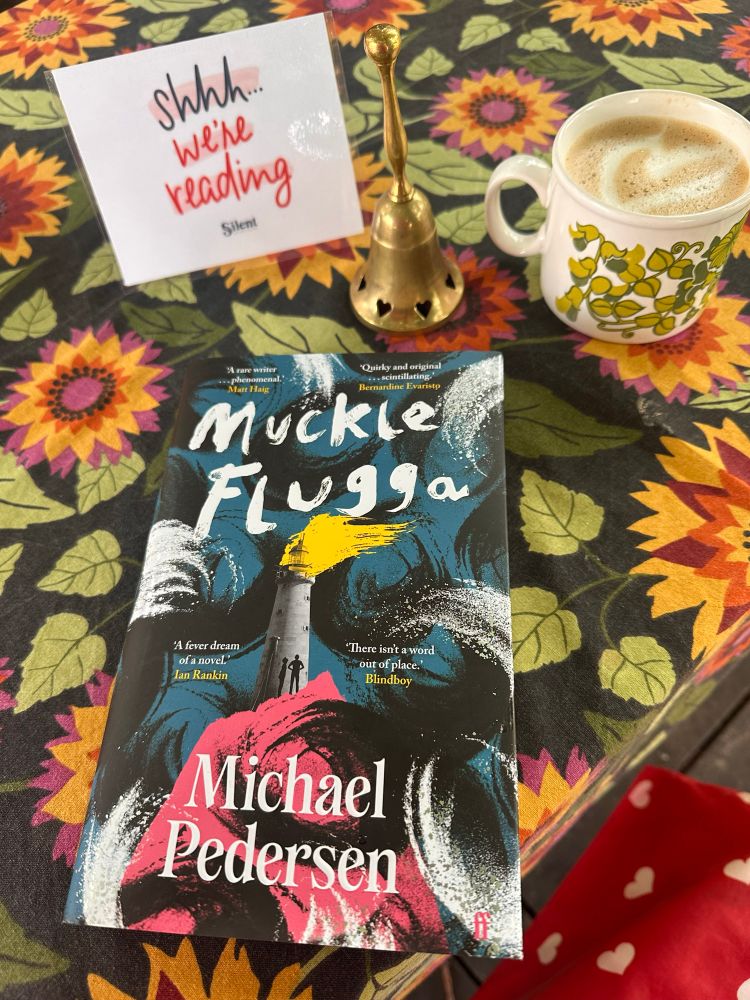 Michael Pedersenin kirja Muckle Flugga pöydällä kukallisen pöytäliinan päällä, Silent Book Clubin kyltti, kahvikupponen ja soittokello vieressään. 
