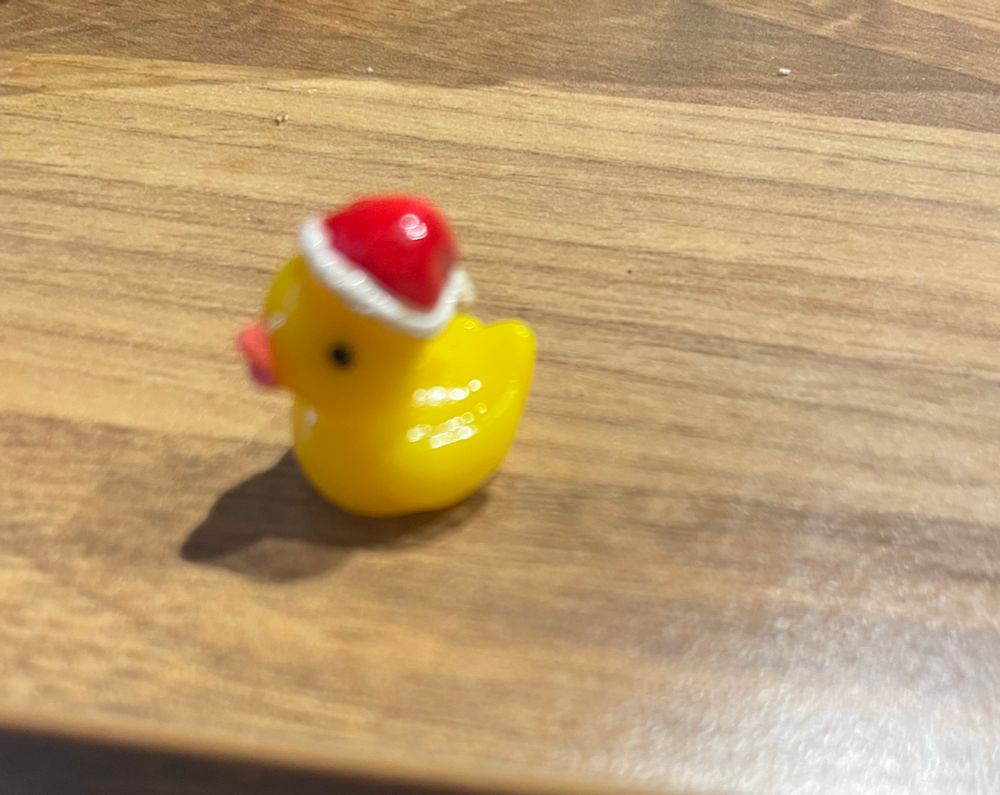 A resin mini duck in yellow with  santa hat on in plastic, on a wooden worktop 