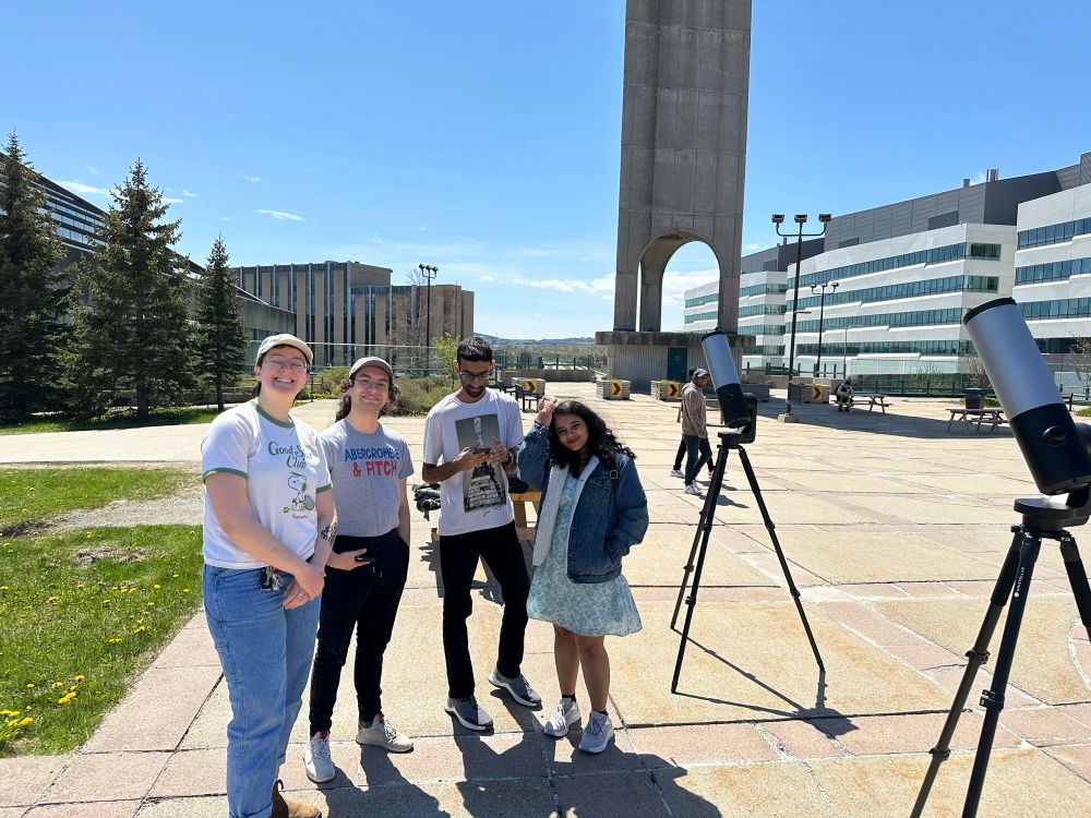 A group of students posing for a picture in front of the Memorial University clock tower next to two telescopes.
