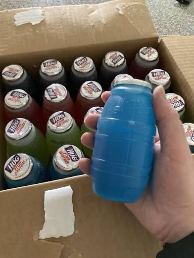 A box of “Little Hug” fruit-flavored drinks in t plastic, barrel-shaped containers with gill lids. A hand in the foreground holds a blue one. No one names the flavors by the actual fruit it’s supposed to mimic; rather, the flavors are just the color of the liquid. And when you’re drinking it and reaching the last sip, it just tastes like plastic. 