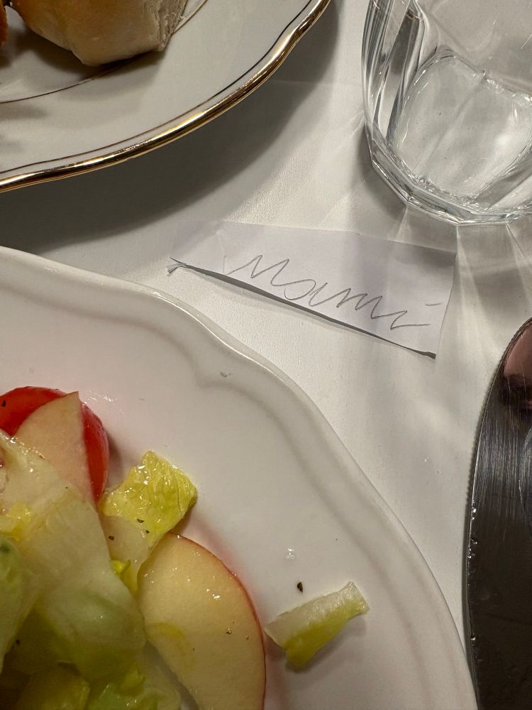 Gedeckter Tisch, im unteren Bildbereich ein Teller mit Salat, rechts daneben ein Messer, über dem Teller ein kleiner handgeschriebener Zettel mit „Mami“ drauf