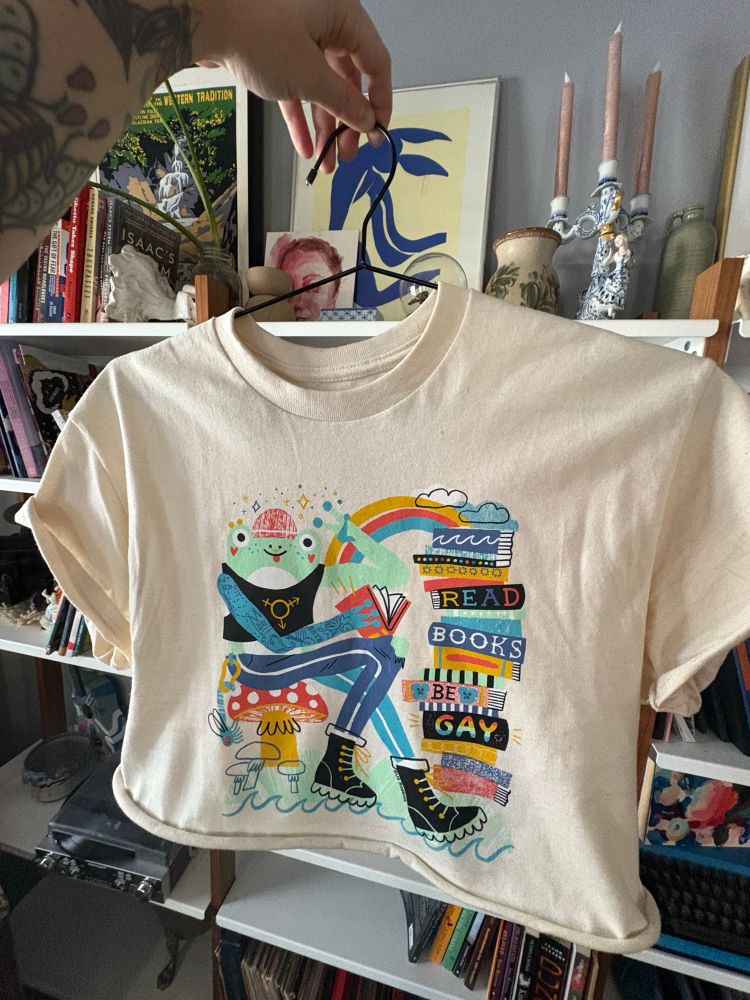 Bug holds up the shirt they designed on a slim black hanger in front of their bookshelf filled with all sorts of art and trinkets. The shirt is a light cream natural color and has a gay punk little frog doing a peace sign and holding a book. They have a beanie, septum piercing, sleeve of frog tattoos, jeans and platform black boots. On their jeans is a caribeaner with a dragonfly key. The frog is sitting on a mushroom on top of a giant lilypad, and to the right is a stack of books as tall as the frog. On the spines of 4 of the books it says “Read Books Be Gay” there is a little rainbow, clouds, and drawn sparkles at the top. Its all drawn in a very cute but printmakery and colorful style.