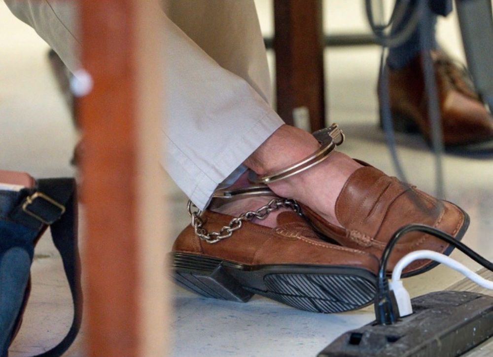 Luigi Mangione’s ankles, demurely crossed, saddle leather loafers on his feet and wearing a pair of khaki chinos. Each ankle is cuffed with a chain dangling down between.