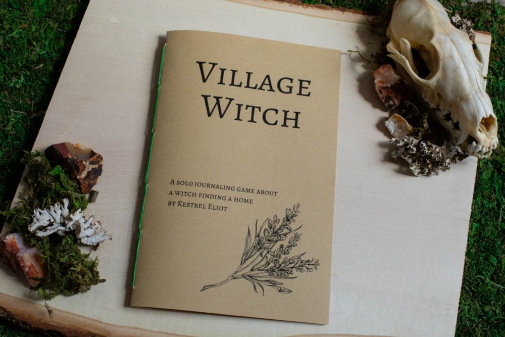 Image of a handbound copy of a book titled Village Witch. It's on a slab of wood surrounded by moss, rocks, and a fox skull