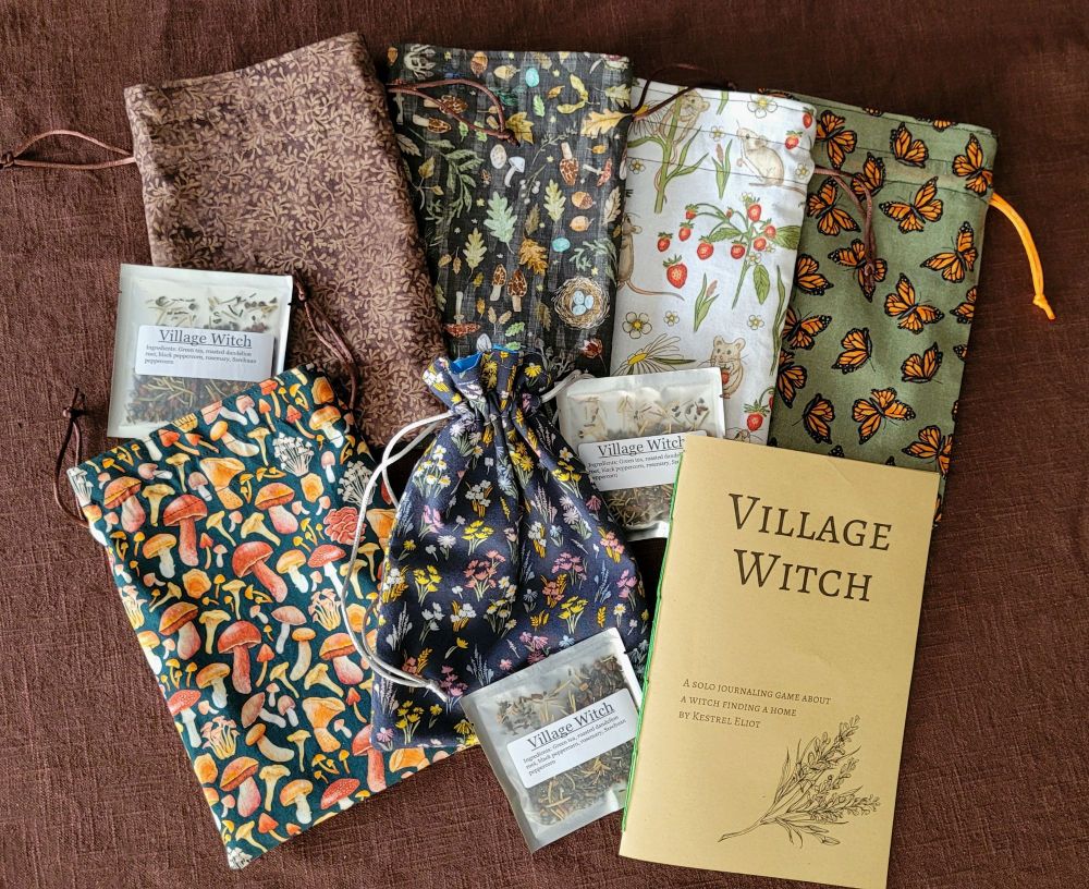 6 handmade tarot bags of varying fabric prints arranged with 3 tea samples and a handbound copy of the solo game Village Witch.