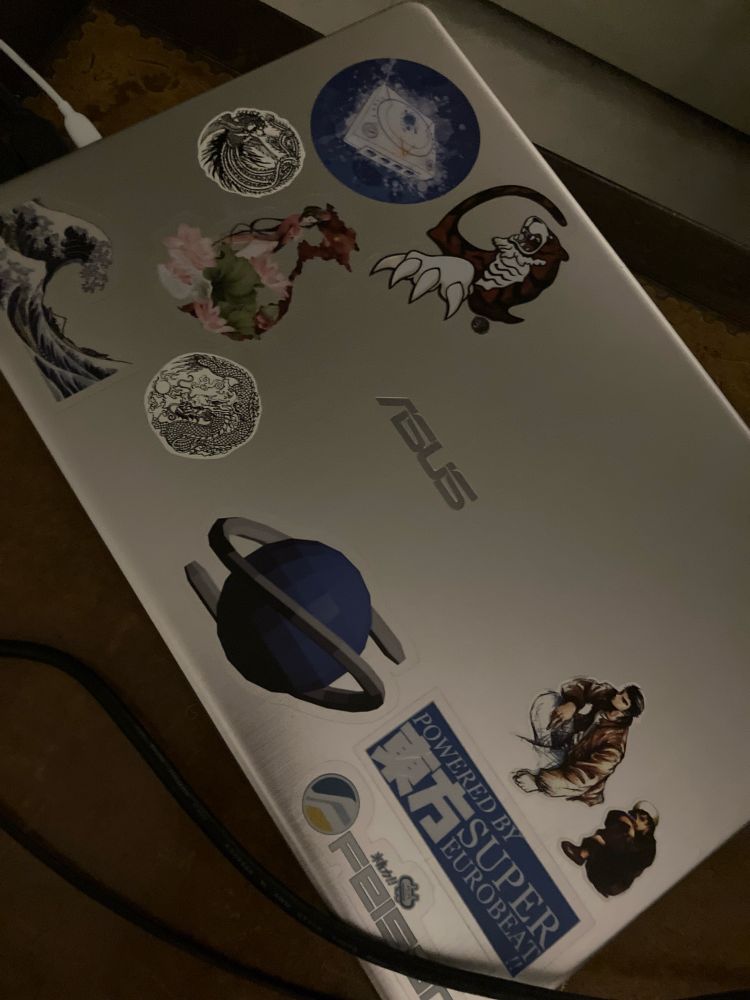 Stickers on top of a grey Asus laptop. They include, from top left: a Dreamcast design; a Hazuki tiger; a phoenix design;  a Vietnamese woman in ao dai next to a map of Vietnam and lotus flowers; a dragon design; the Great Wave off Kanagawa; a Sega Saturn design; Ryo and Shenhua from Shenmue; a sticker saying “Powered by 東方 Super Eurobeat!!”; and the logo and wordmark for the Feisar team from Wipeout.
