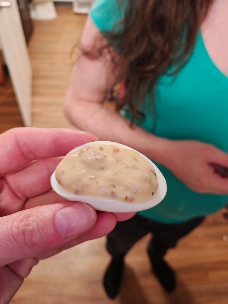 A woman's hand holding a deviled egg in a kitchen with a hardwood floor as another woman with long, brown hair and a green shirt stands by (she is waiting to eat the egg)