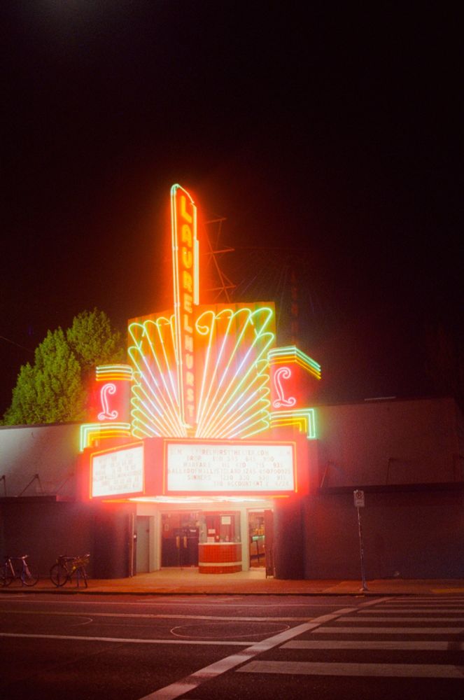 A photo of the front of a theater at night, the massive neon display of it's name, "Laurelhurst" glowing in the night. Two bicycles are chained up on racks out front, and a tree is visible behind the building and signage.