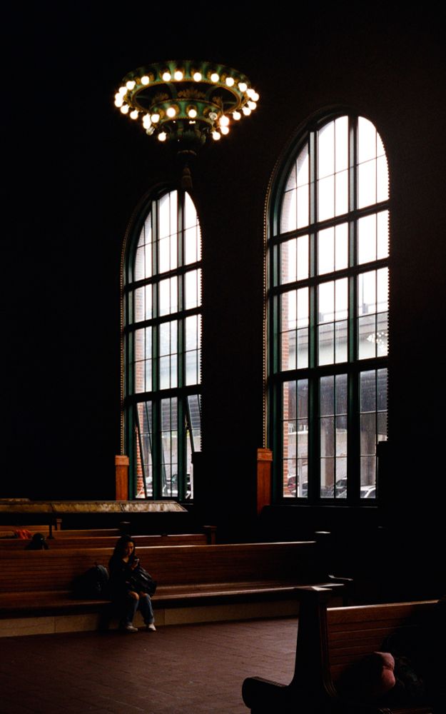 A color photo of the inside of a train station on a rainy day. A woman sits on a large, empty wooden bench, looking at her phone, while a pool of light from outside has illuminated her and the smooth brick floor beneath her. Above her is a large wrought iron chandelier, and in the background are huge windows looking out onto an elevated road.