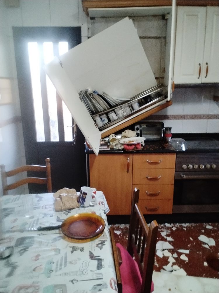 A kitchen cabinet that has fallen over, the floor is filled with broken plates and mugs.