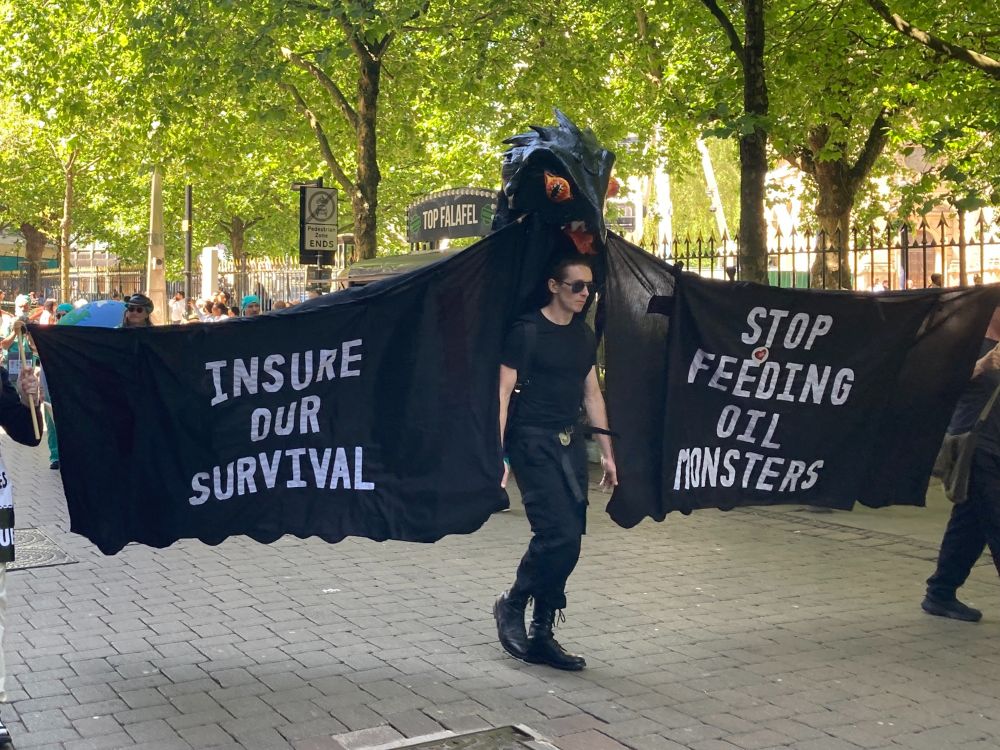 Oily Monster: A man dressed in a black carries a  slick, shiny monster puppet, banners either side read 'Insure Our Survival' and 'Stop feeding oil monsters'
