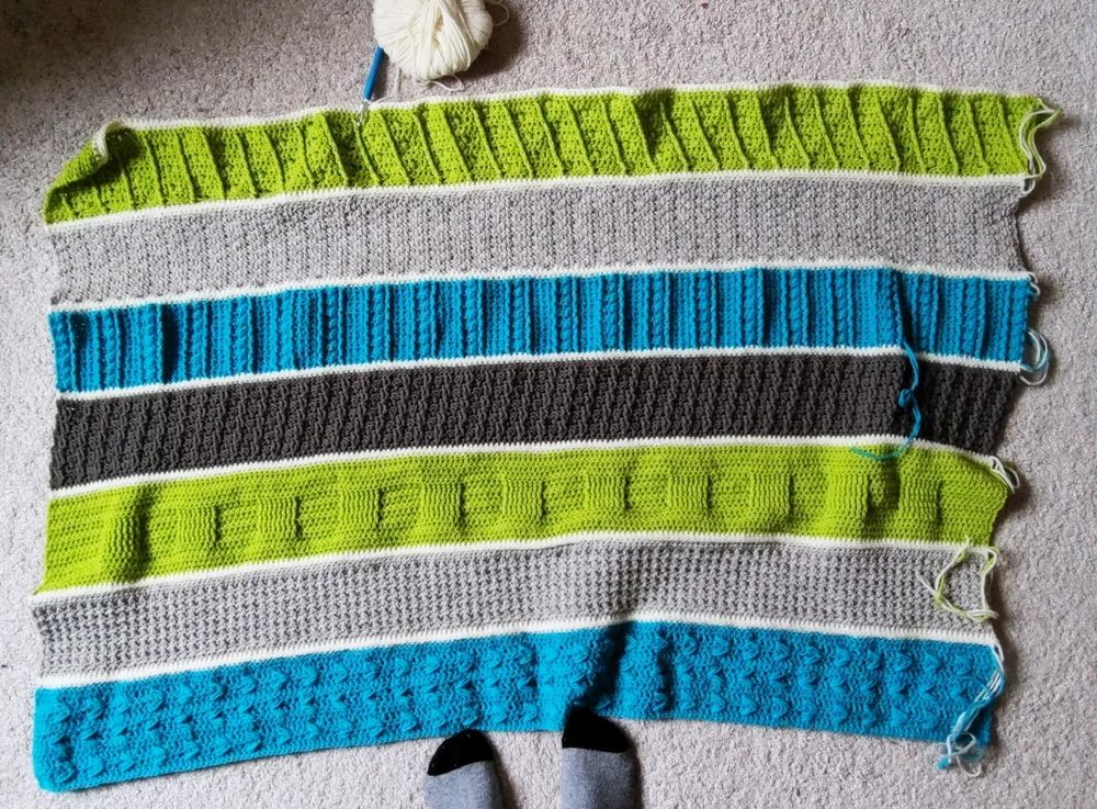 An unfinished crocheted blanket, with loose ends and an attached ball of yarn, laid out on the floor. It has 7 stripes in robin's-egg blue, light brown, chartreuse, and dark brown, repeating, with a thin bar of cream separating each stripe. Every stripe has a different texture (some have ridges, puff stitches, squares set out in relief, etc.). There are gray-socked toes poking into the bottom of the picture for scale (it's about four feet wide and three feet long).