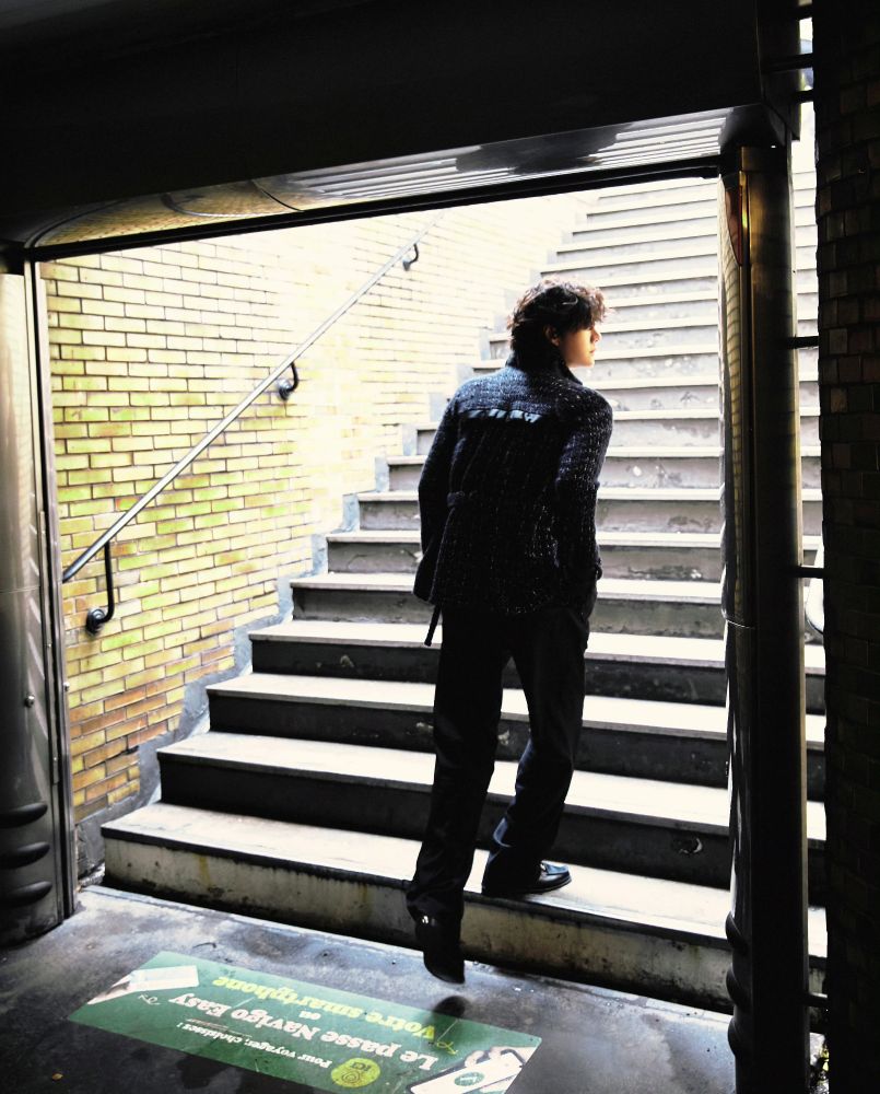 Wang Yibo with a head of soft curly hair, in a dark glittery Chanel outfit, heading up the stairs out of the Paris subway into the sunlight, his face turned to the right and glowing gently in the light.