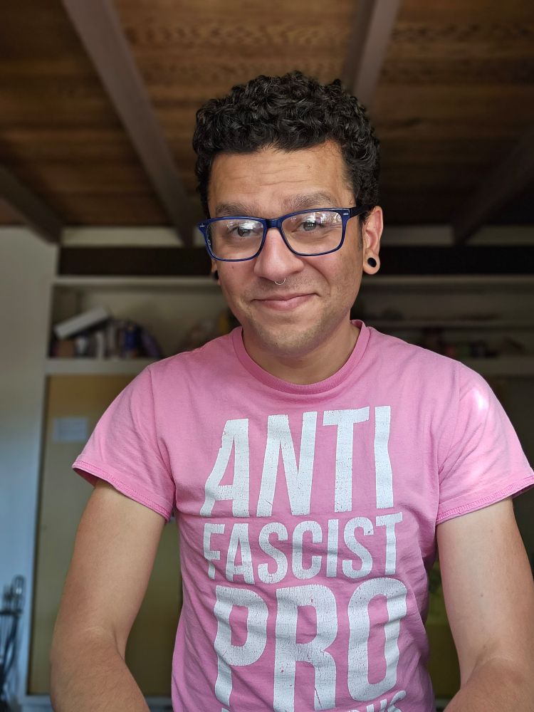 Jeremy, a person with curly dark hair, blue framed glasses, blue glass 0g plugs in their stretched earlobes, and a pink shirt reading "Antifascist Pro Fabulous". 
