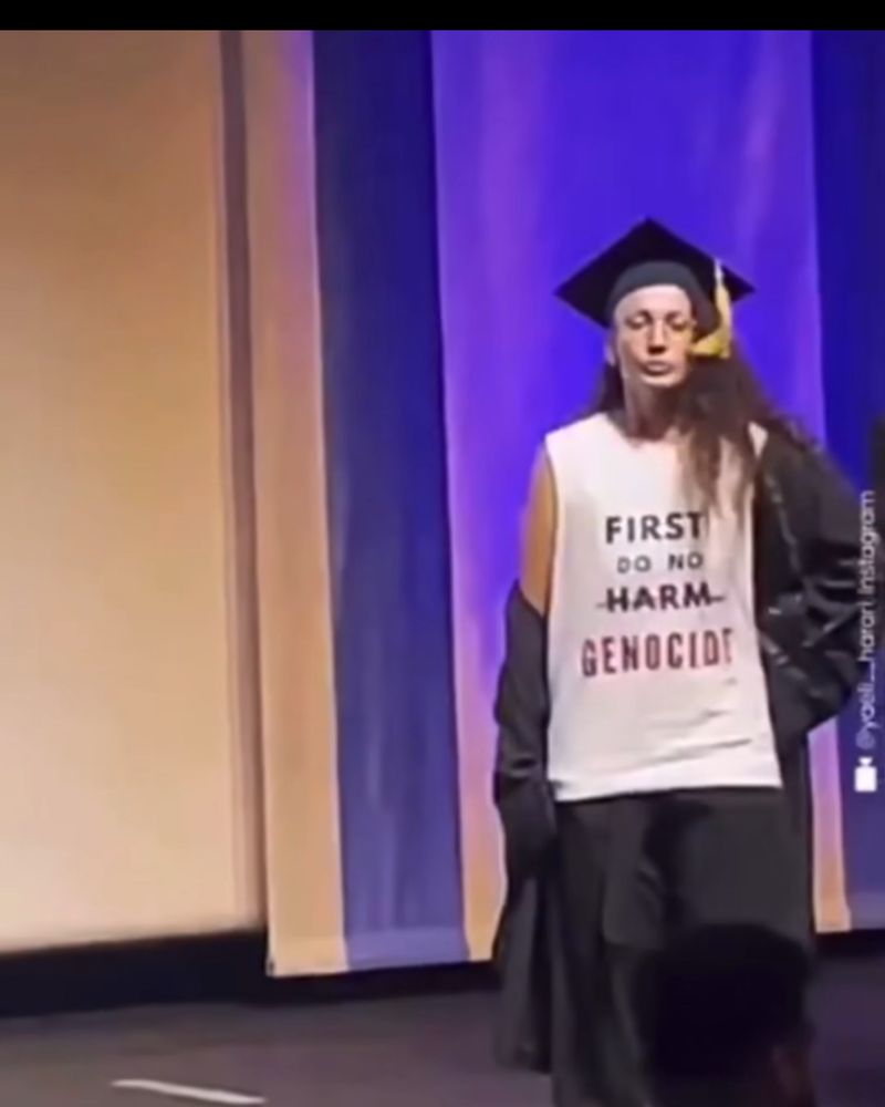Person in cap and gown. TShirt says: first do no harm, and harm is crossed out and it says GENOCIDE 