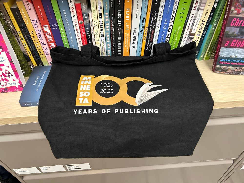 A black tote bag printed with the golden colored Centennial logo for the University of Minnesota Press. It says 100 years of publishing 1925-2025. 