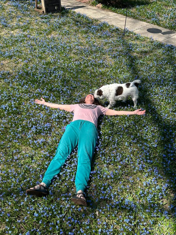 Me laying down in my backyard which is covered in tiny blue flowers, squill. My dog Mr. Bill, who is white with brown spots, is sniffing my face. My arms are outstretched in the sunshine. The world is on fire but it’s my birthday and I’m trying to feel ok. It’s working. 