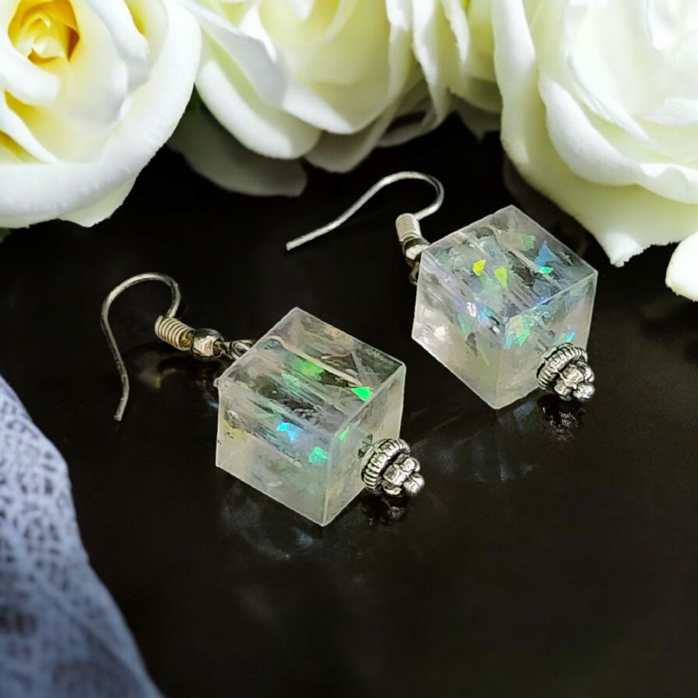 A pair of clear resin cube earrings with colorful inclusions on a dark counter top with white roses.