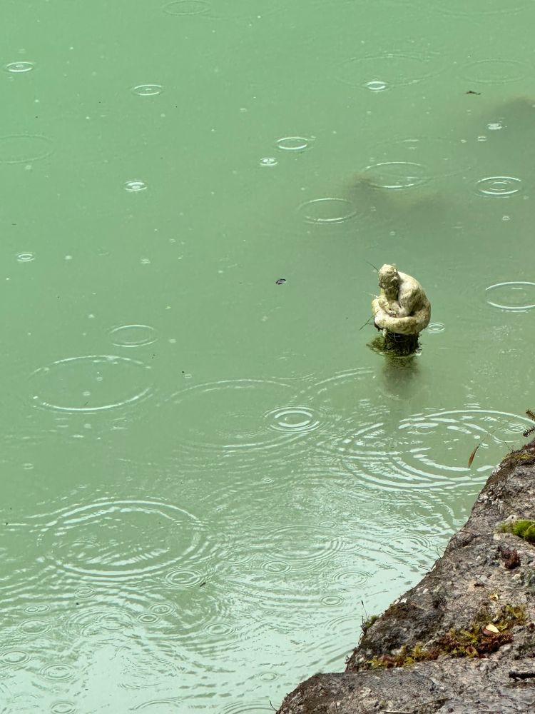 Small statue in a clear green-turquoise pond at Wheal Martyn Clay Works. Rain drop ripples in the water