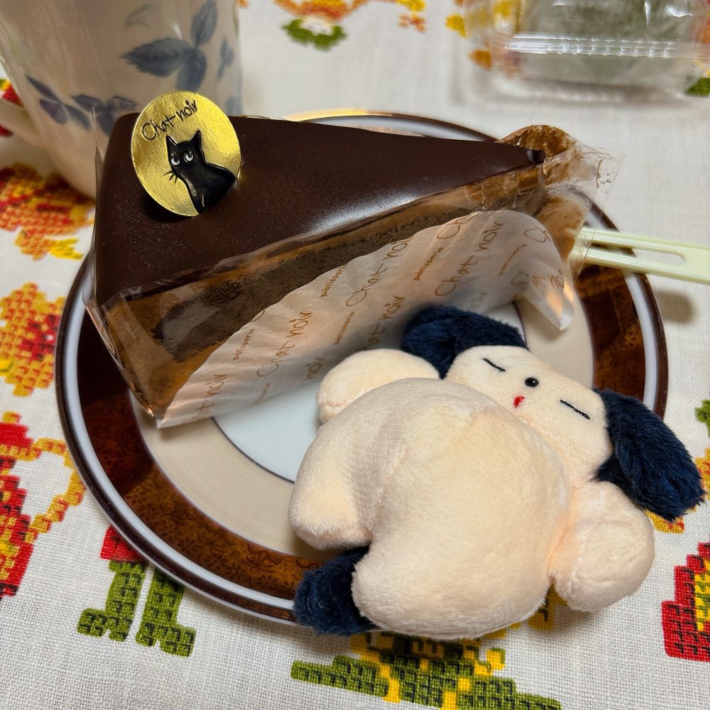 チョコレートケーキの写真。金色の丸いシートに黒猫と”Chat noir”と黒字で描かれた飾りがついている。お腹のふっくらとした犬のぬいぐるみを添えて。