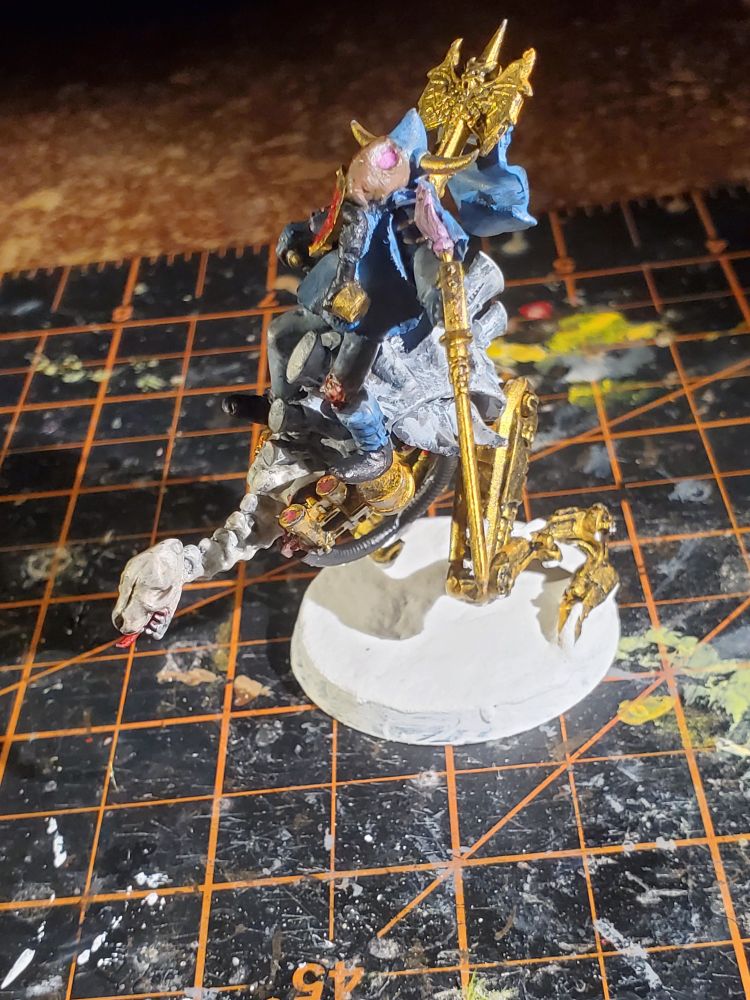 Kitbashed miniature model with a swordsman in blue clothes and gas mask carrying a sword while mounted around an eyeless cyborg dog monster with mechanical legs, on a snowy base.