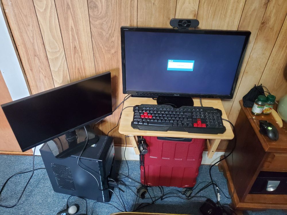 Computer monitors precariously perched on a wooden tray and suitcase, with a second monitor overhanging and placed on the computer tower itself. Various wires are running all over each other due to lack of proper desk.