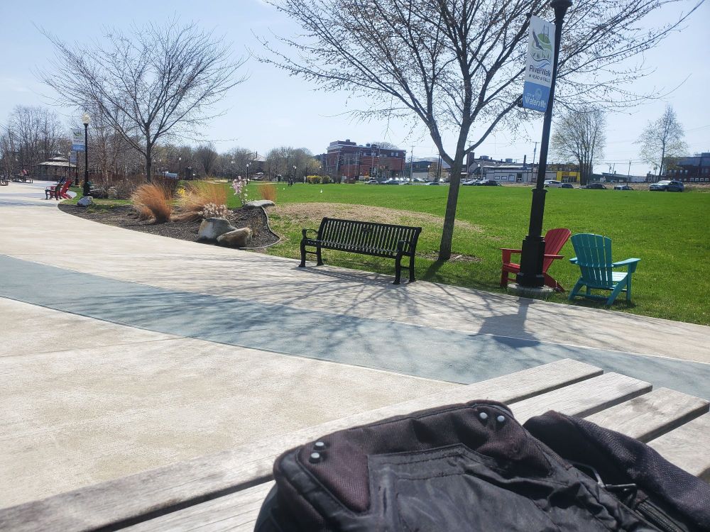 Sidewalk in a green park with a few trees and benches and chairs. It is a clear day.
