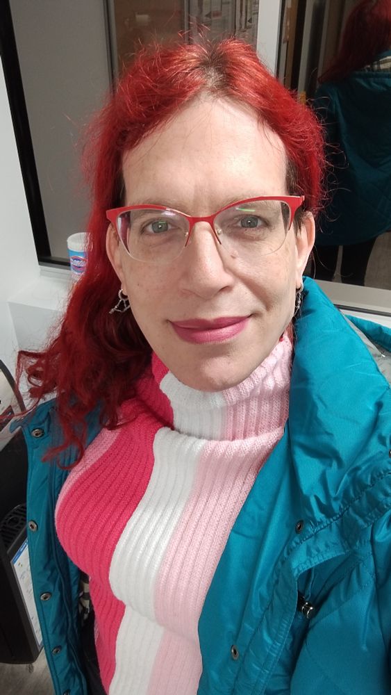 Ashley wearing a teal puffy jacket and a striped sweater. The sweater has wide vertical stripes of white and different shades of pink, resembling part of the lesbian pride flag.