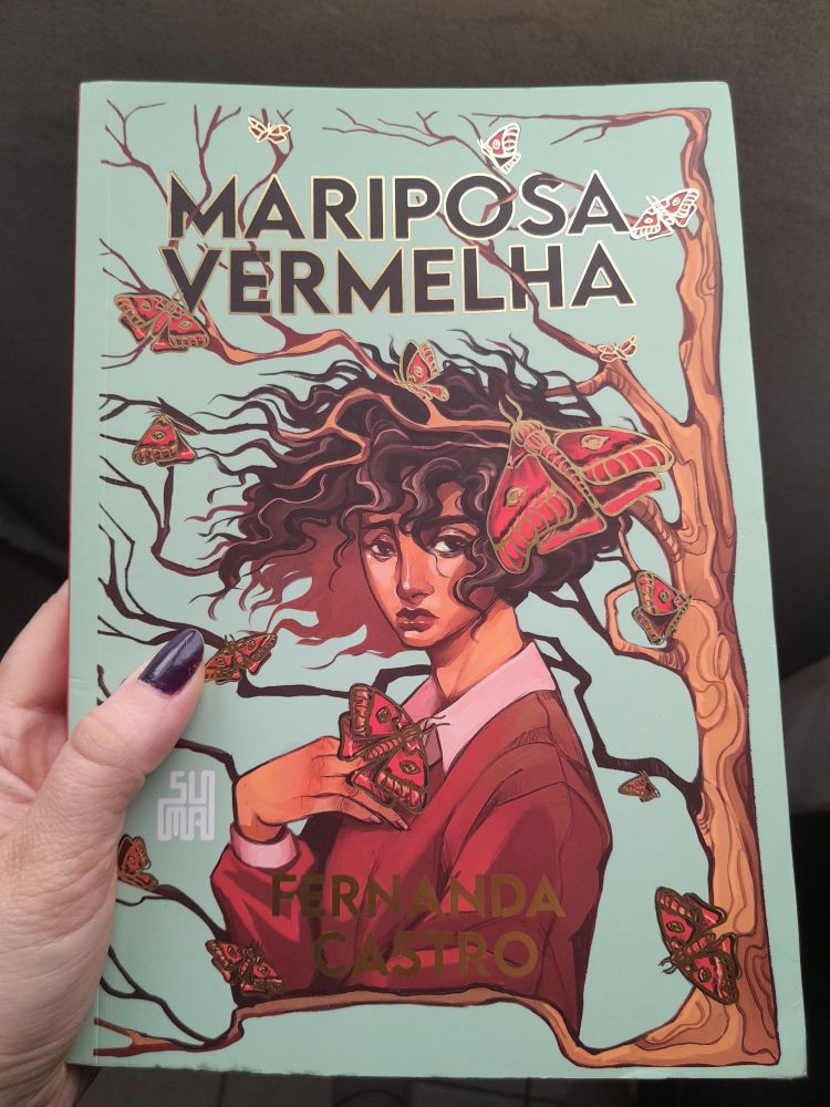 Capa do livro Mariposa Vermelha. O fundo é verde claro, com a ilustração de uma garota de cabelos cacheados esvoaçantes e pele negra clara. Ela está sob uma árvore de galhos secos cujo tronco ocupa a lateral direita da capa, seus cabelos se confundem com os galhos que atravessam a capa. Espalhadas, mariposas vermelhas com contorno dourado. A garota tem um semblante triste e está vestida com uma camisa social beanca e um pulôver marrom claro. O título vem no topo, em marrom bem escuro e contornos dourados nas letras. O nome da autora Fernanda Castro está na parte debaixo em letras douradas. Uma mão branca segura o livro do lado esquerdo. 