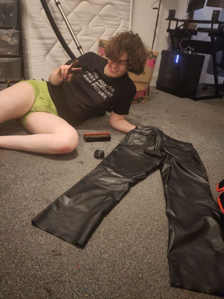 yvette laying next to leather trouser xe is polishing for some reason? xyr shirt reads: "YOU ARE THE PAST PRESENT AND FUTURE". it's a jenny holzer one.