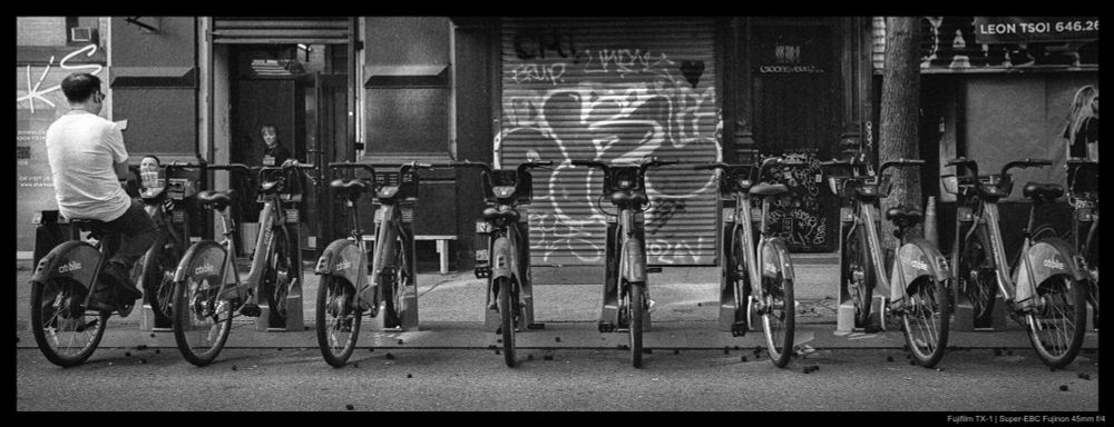 A man sits atop a docked Citibike, with many similarly docked bicycles running through the frame from left to right.  Shuttered storefronts are visible in the background.