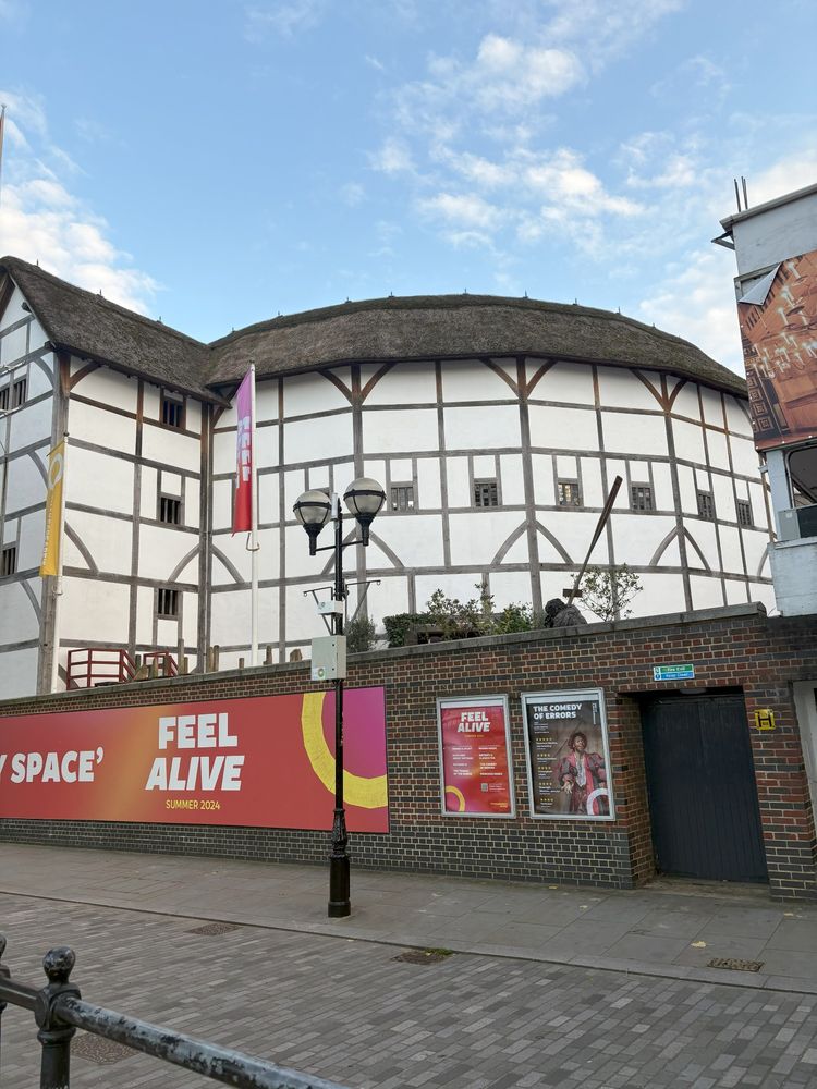 The exterior of the Globe Theatre in London, England. White building with brown trim with a brick wall in front of it.