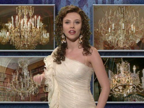 A screenshot from a Saturday Night Live infomercial. Scarlett Johansson wears a one-shouldered cream gown and stands in front of a backdrop featuring four large photos of ornate crystal chandeliers. She has curly brown hair and wears large dangling earrings, with her mouth slightly open as if speaking or singing. The image has a formal, staged feel, resembling a television or performance set.