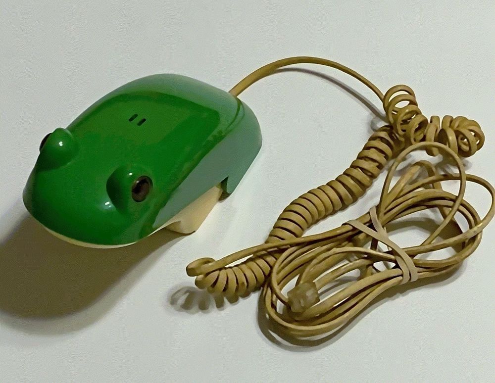 A vintage novelty corded telephone designed to look like a green frog. The phone's receiver resembles a frog's head and body, complete with raised eyes and a smooth, rounded shape. The beige coiled cord is attached and bundled with a rubber band, ending in a clear RJ11 connector. The phone rests on a plain white surface.