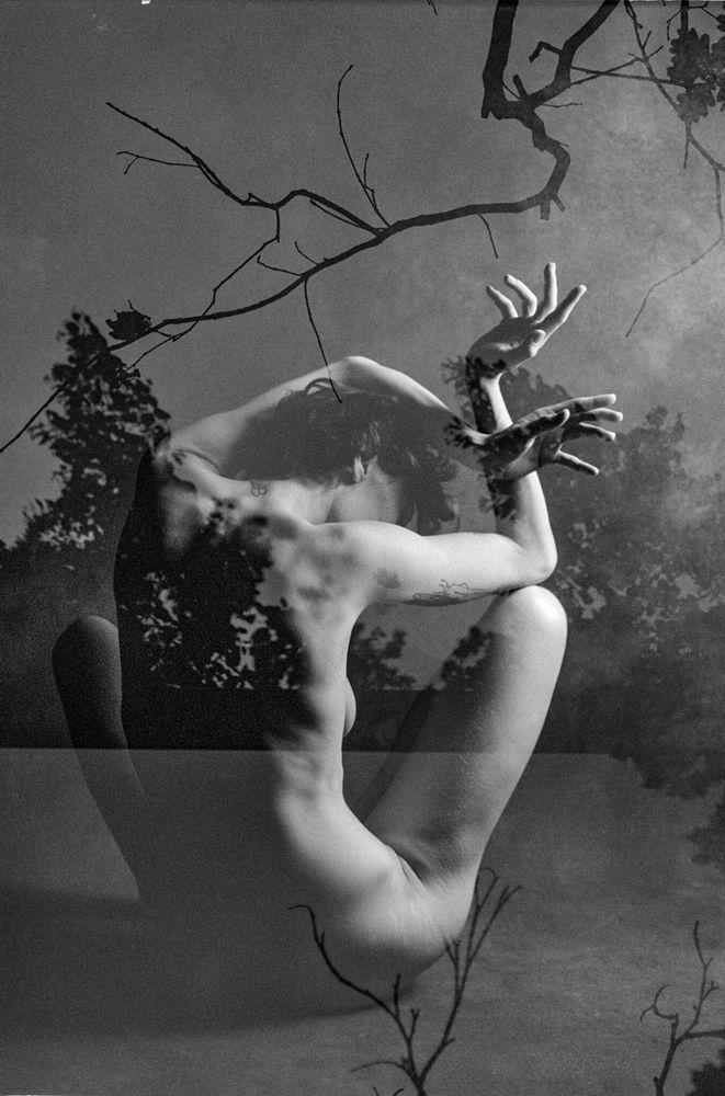 Analoge Doppelbelichtung in schwarz-weiß, Nude art, sitzende, ihre Hände nach oben haltende Frau, umgeben von Ästen