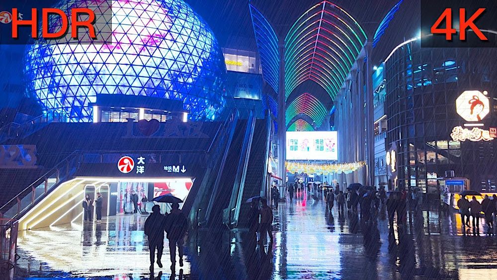 Bright picture of a Chinese mall at night, labeled "HDR" and "4K." Has a brilliant blue light globe on the left and a walkway under it, and another walkway through the middle with green and red arched ribs over it, and a black mirrored and rounded building on the right. People walk huddled under umbrellas as it's raining.