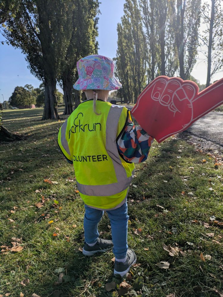 Toddler stood in a tree lined path wearing a high-vis jacket and holding a large, red, foam hand