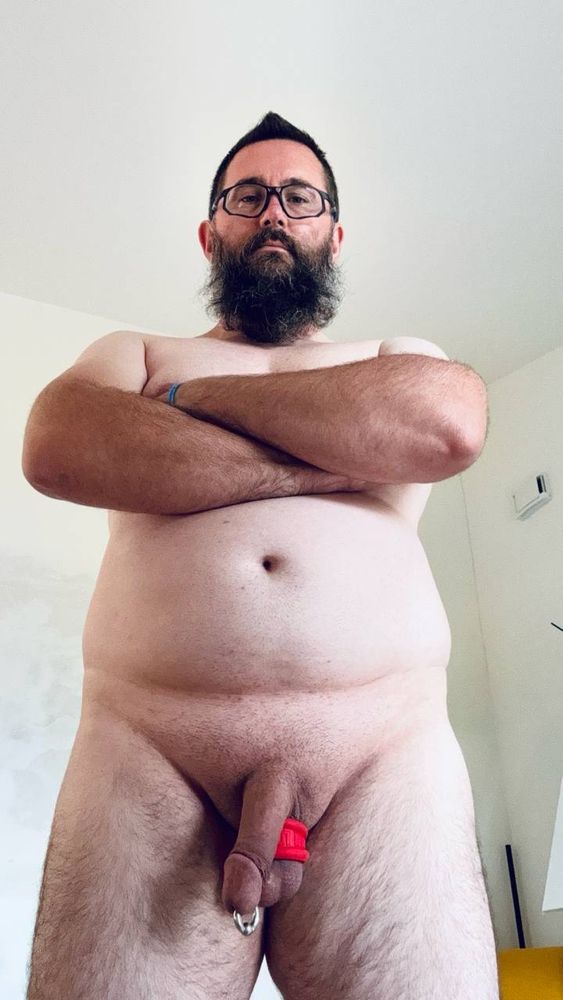 Adult male subject standing in a frontal position, photographed from a low angle. Fully nude body with exposed genital area; circumcised. Features a Prince Albert piercing and a silicone ball stretcher applied at the base of the scrotum for testicular traction. The pose is firm, with arms crossed over the chest and gaze directed at the camera. The composition evokes a symbolic reading of the body as a conscious, modified space, aligning with the aesthetics and philosophy of the Modern Primitives movement.